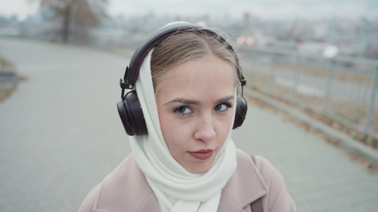 自信を持ってヘッドホンを調整するエレガントな女性。街のスカイライン近くでヘッドホンと共にポーズをとるファッショナブルな女性。編み込みヘアとスタイリッシュなコートを身につけ、アップビートな音楽が流れる都会の環境にいるシックな女性。