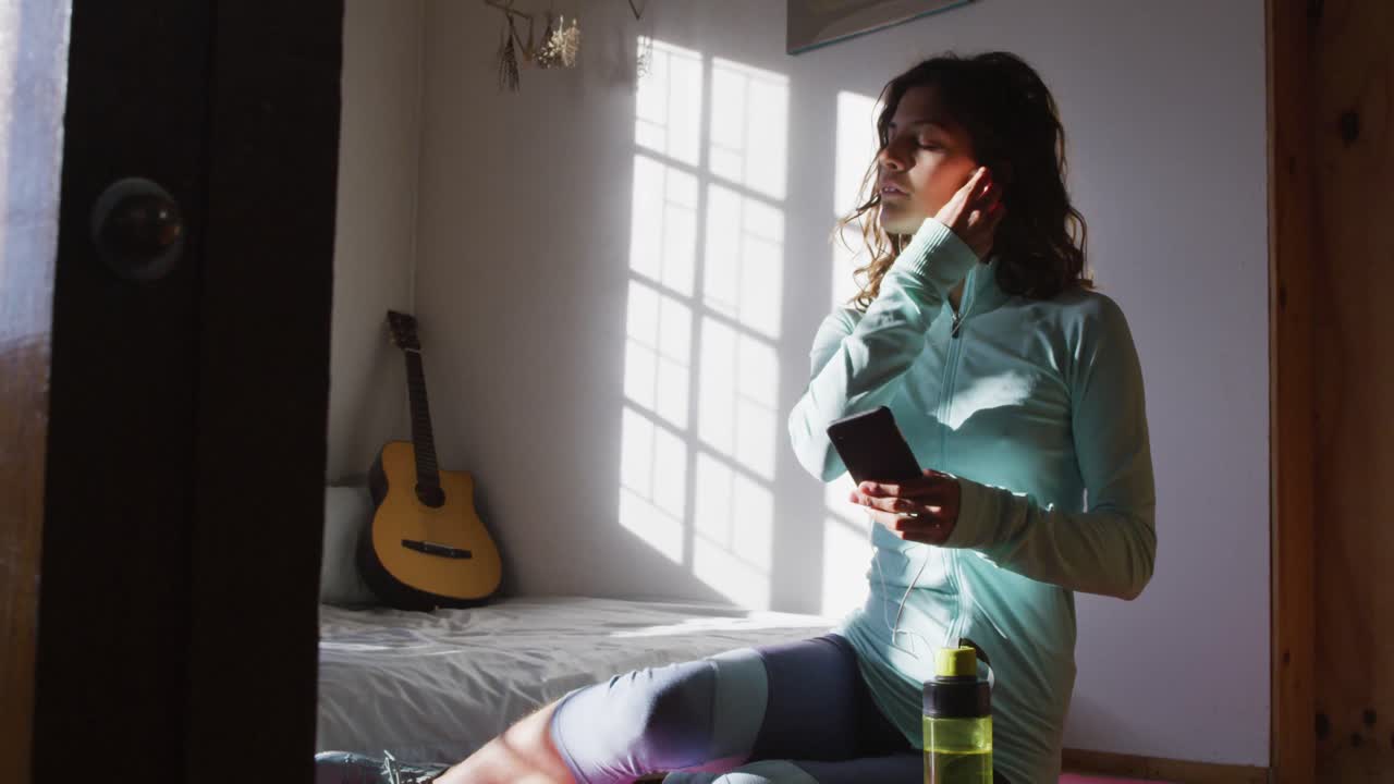 mujer de raza mixta feliz preparándose para el entrenamiento, sosteniendo un teléfono inteligente poniendo auriculares en una habitación soleada