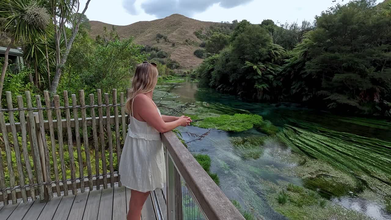 Uma mulher está em uma plataforma de observação admirando as águas cristalinas e os arredores exuberantes de Blue Springs, na Nova Zelândia. O cenário sereno destaca a beleza natural da área.