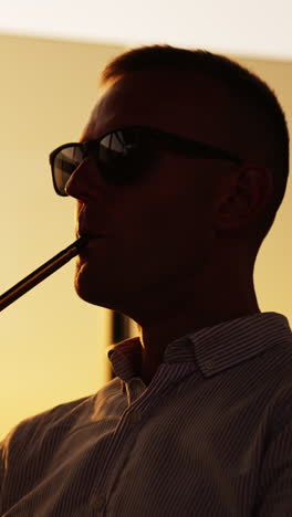man in dark glasses sitting at sunset holding hookah pipe. relaxing male making thick smoke from nargileh. close up. vertical video