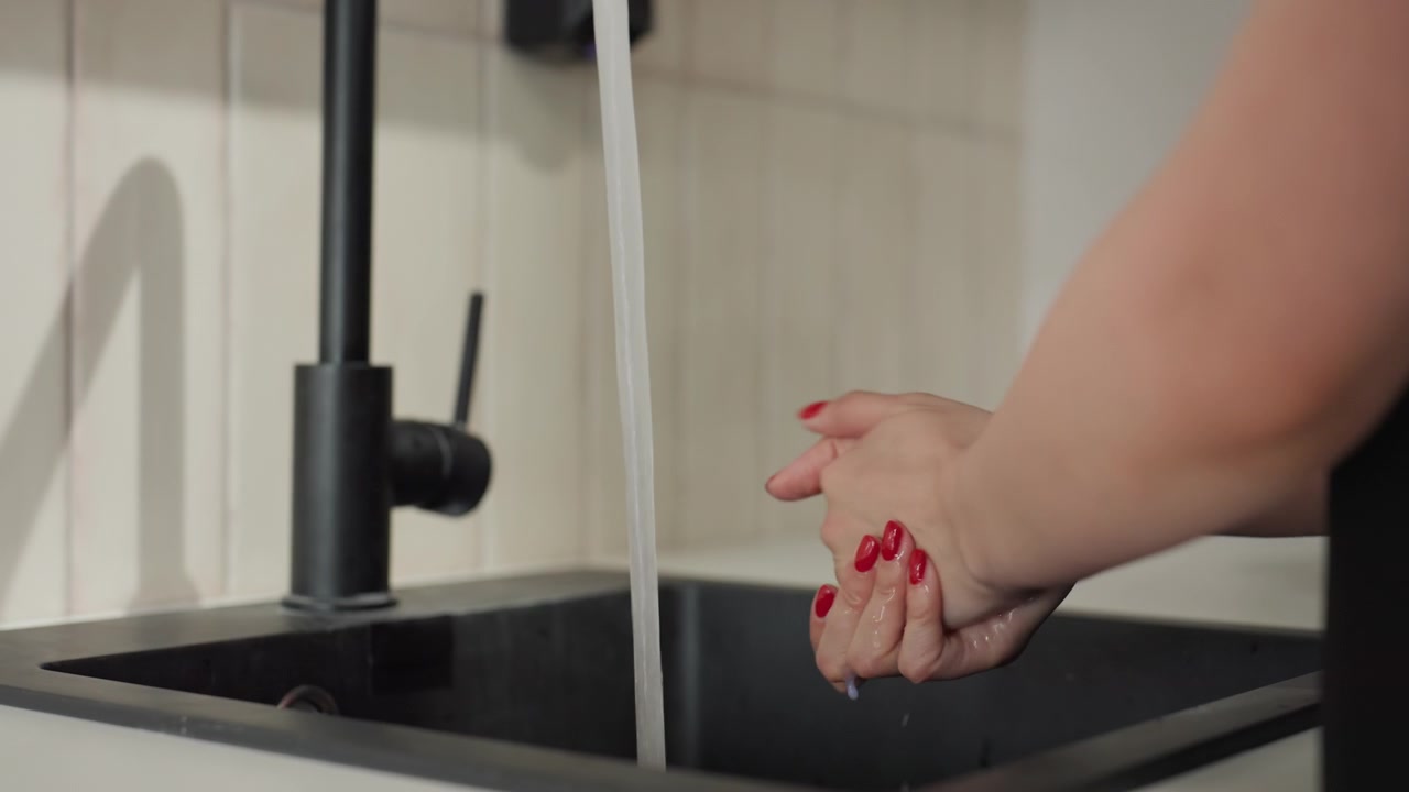 Primo piano di mani che si lavano sotto acqua corrente in un moderno lavello da cucina. La persona manipola il rubinetto per controllare il flusso mentre pulisce accuratamente la pelle con smalto rosso visibile.