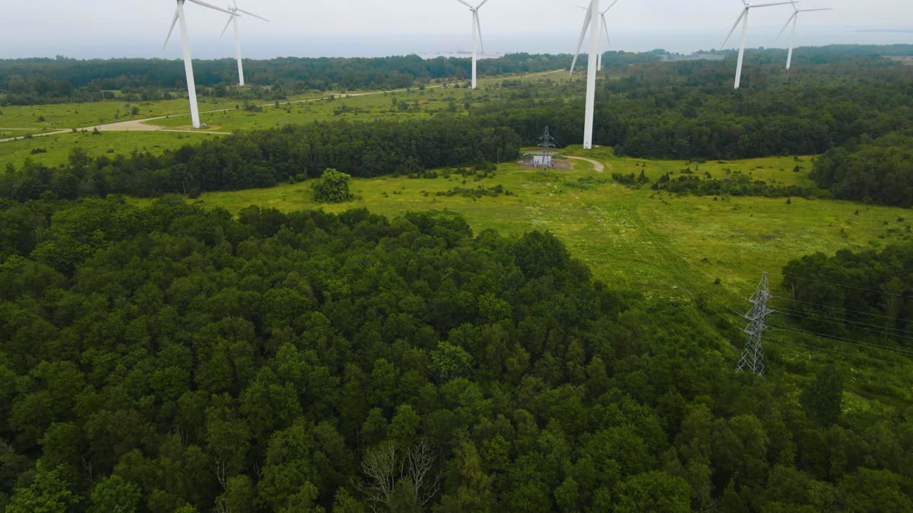 Aerial drone revealing footage shot of large white renewable energy electrical power creating wind turbines on a green forest covered peninsula during a cloudy day. Footage flying backwards smoothly