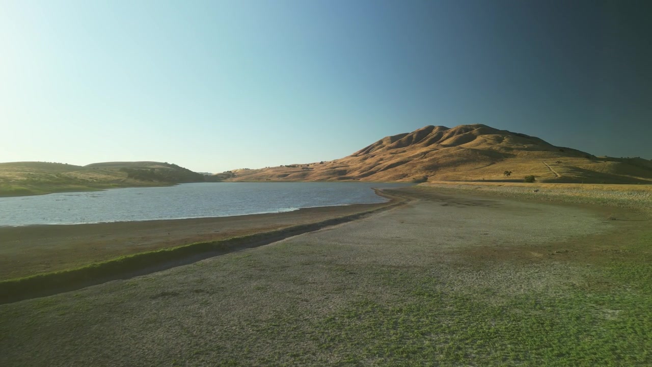 A steady dolly zoom in above the Paicines Reservoir, the drone captures the peaceful water surface meeting rural farmland and clear blue skies.