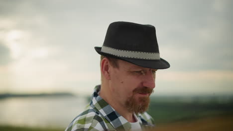 close-up of a man wearing a black hat and a green checkered shirt, deeply focused on an unseen task. the background features a serene, out-of-focus landscape