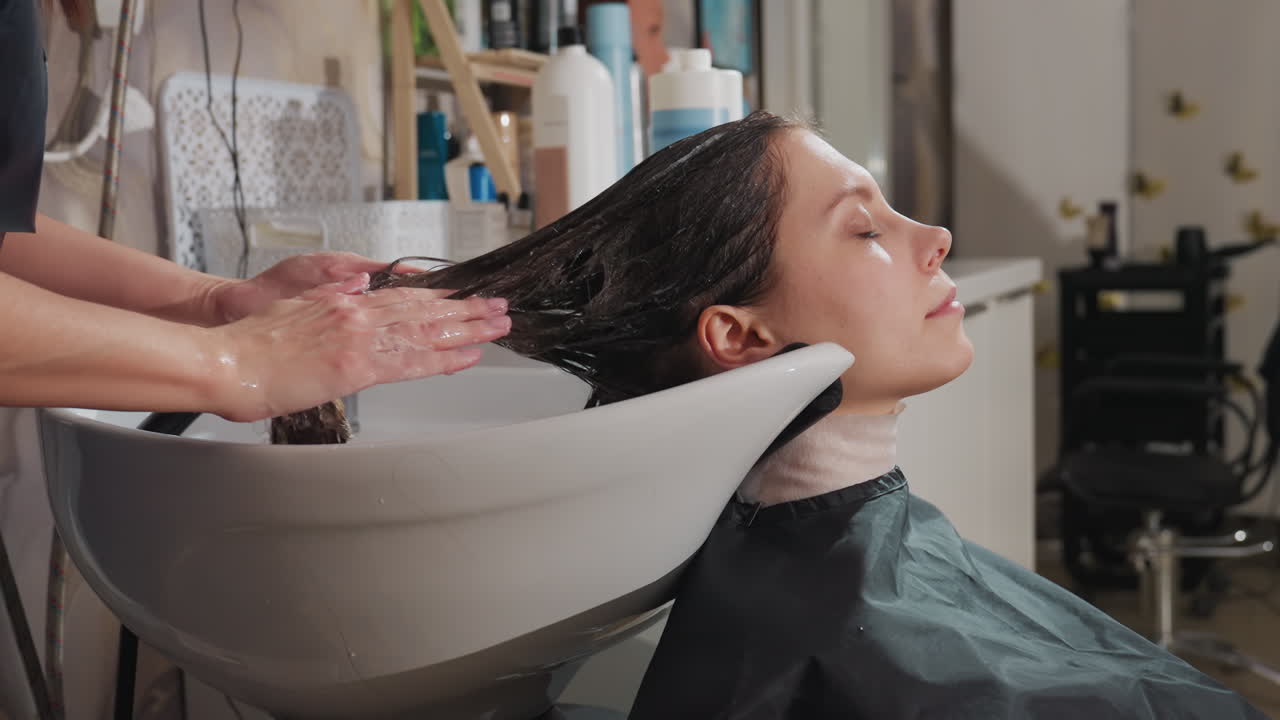 헤어 케어 서비스를 받는 동안 차분한 분위기, 미용실에서 편안하게 샴푸와 마사지를 즐기는 고객, 스타일리스트가 진정 효과가 있는 두피 마사지와 꼼꼼한 머리 감기를 제공하는 평화로운 환경