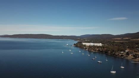 Aerial-drone-pan-around-collection-of-boats-in-deep-blue-sea-surrounded-by-small-mountain-range-and-large-factory-building-on-clear-sunny-day