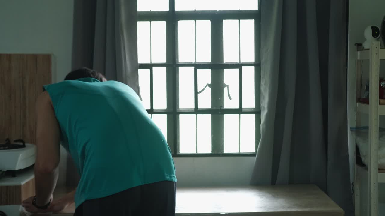 Static wide shot of young man wiping and cleaning kitchen table by sunlit window.
