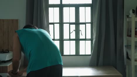 static wide shot of young man wiping and cleaning kitchen table by sunlit window.