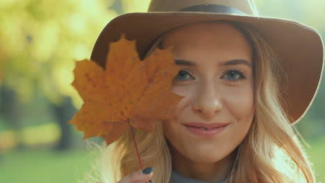Close-up-view-of-Caucasian-young-blonde-woman-wearing-a-hat-smiling-and-hiding-her-face-behind-a-yellow-autumn-leaf-in-the-park
