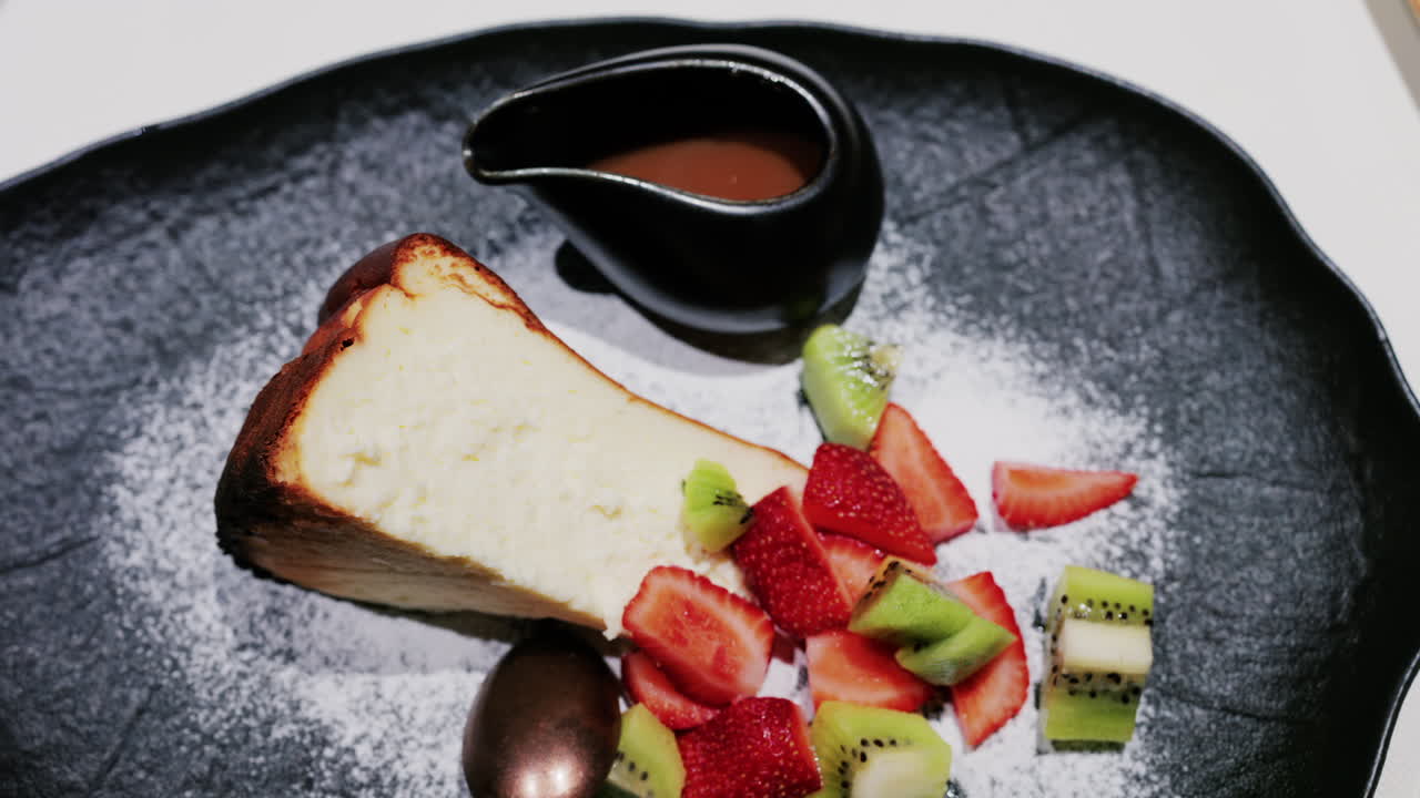 A slice of cheesecake served with fresh strawberries and kiwi, garnished with powdered sugar on a plate at a restaurant