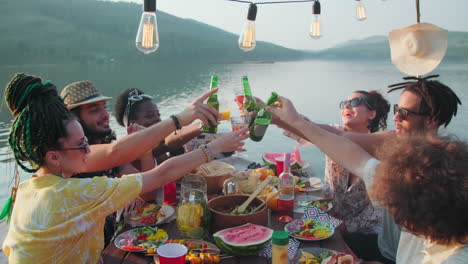 young people clinking glasses in toast at dinner party by lake