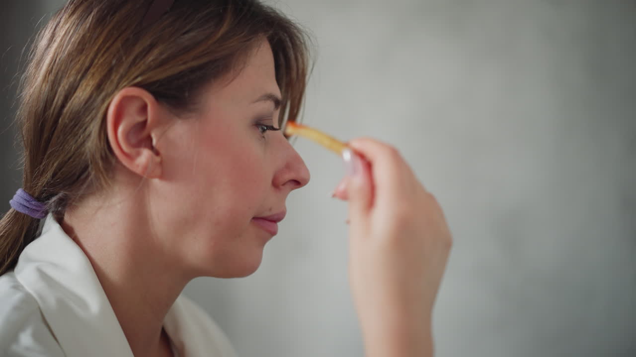 Side view of lady wearing white eating potato chip dipped into red sauce focused on something unseen with hair tied back in ponytail sitting calmly against light background