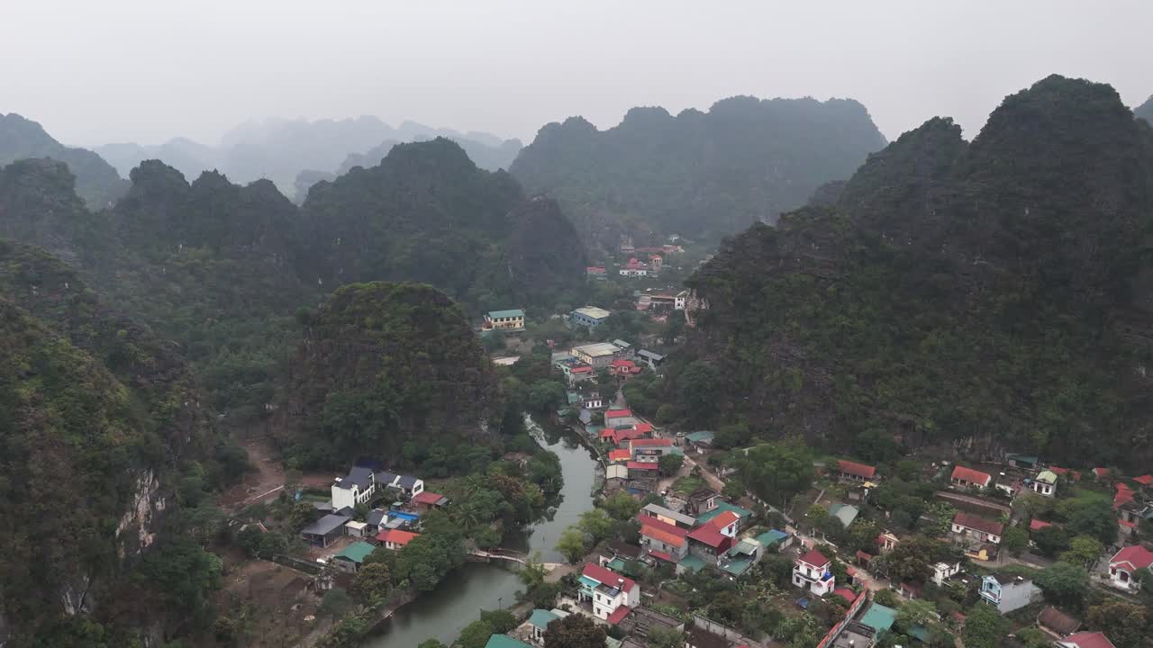 Aerial view of Trang An in Ninh Binh, Vietnam, featuring a winding river, colorful village houses, and dramatic limestone karst mountains surrounded by lush green landscapes under a hazy sky