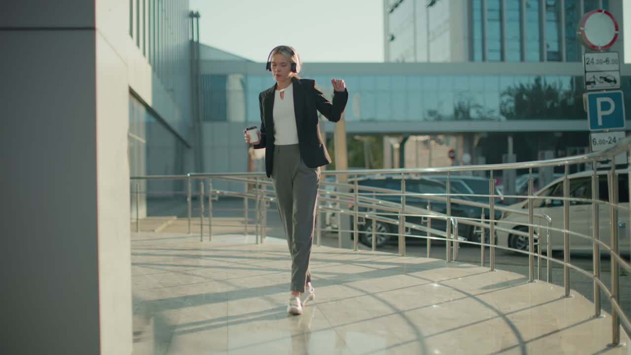 Woman holding beverage dancing joyfully to music outdoors with parked cars and another car driving out in background near glass office buildings and metallic railings in bright morning light