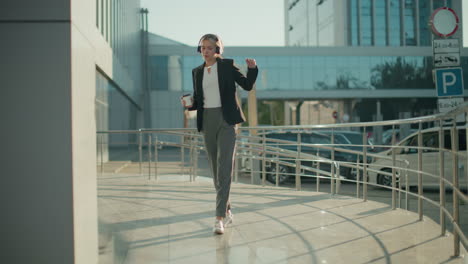 woman holding beverage dancing joyfully to music outdoors with parked cars and another car driving out in background near glass office buildings and metallic railings in bright morning light