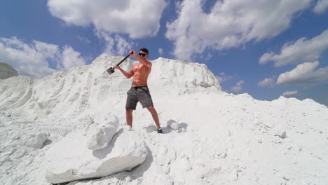Muscle athlete strongman in a cap and sunglasses man hits a huge white rock with a hammer in the white sandy mountains or in a quarry. slow motion. sunny day . White landscape.