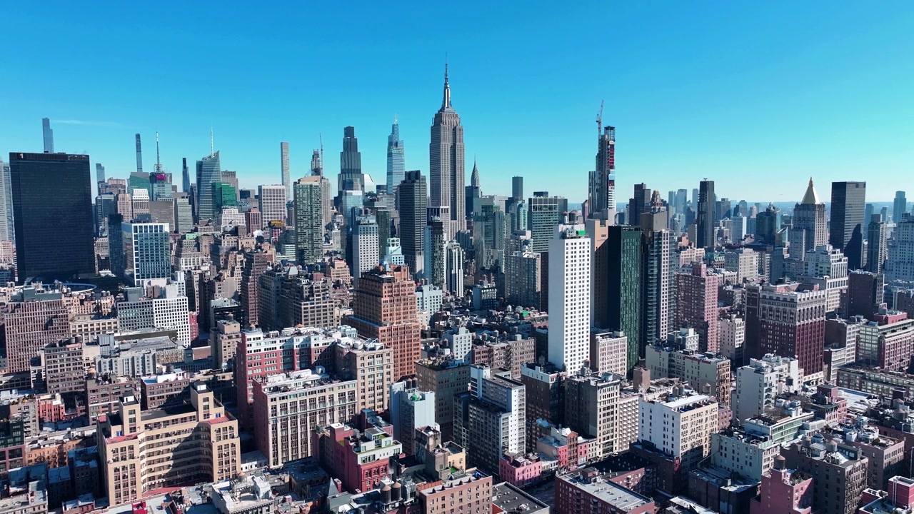 Stunning high-angle aerial view of iconic New York City skyline on a bright sunny day. Empire State Building towers over dense Manhattan skyscrapers under clear blue skies.