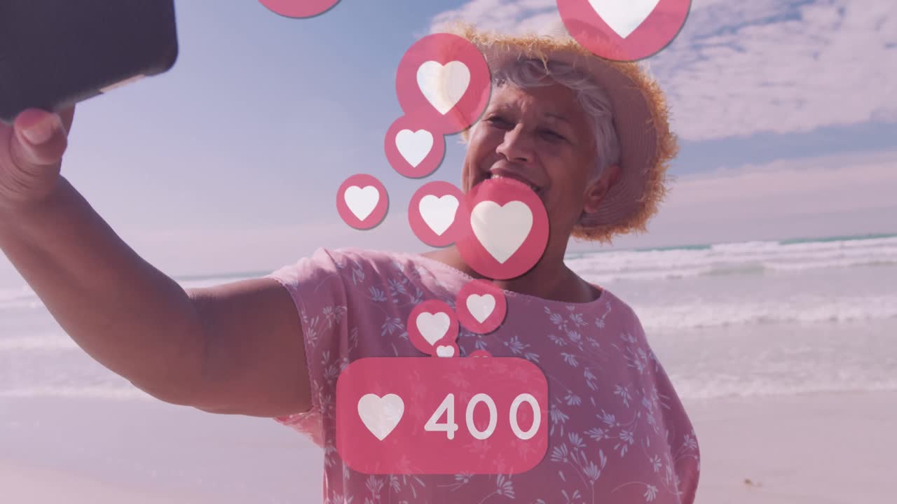 animación de corazones sobre feliz anciana mujer afroamericana tomando una selfie en una playa soleada