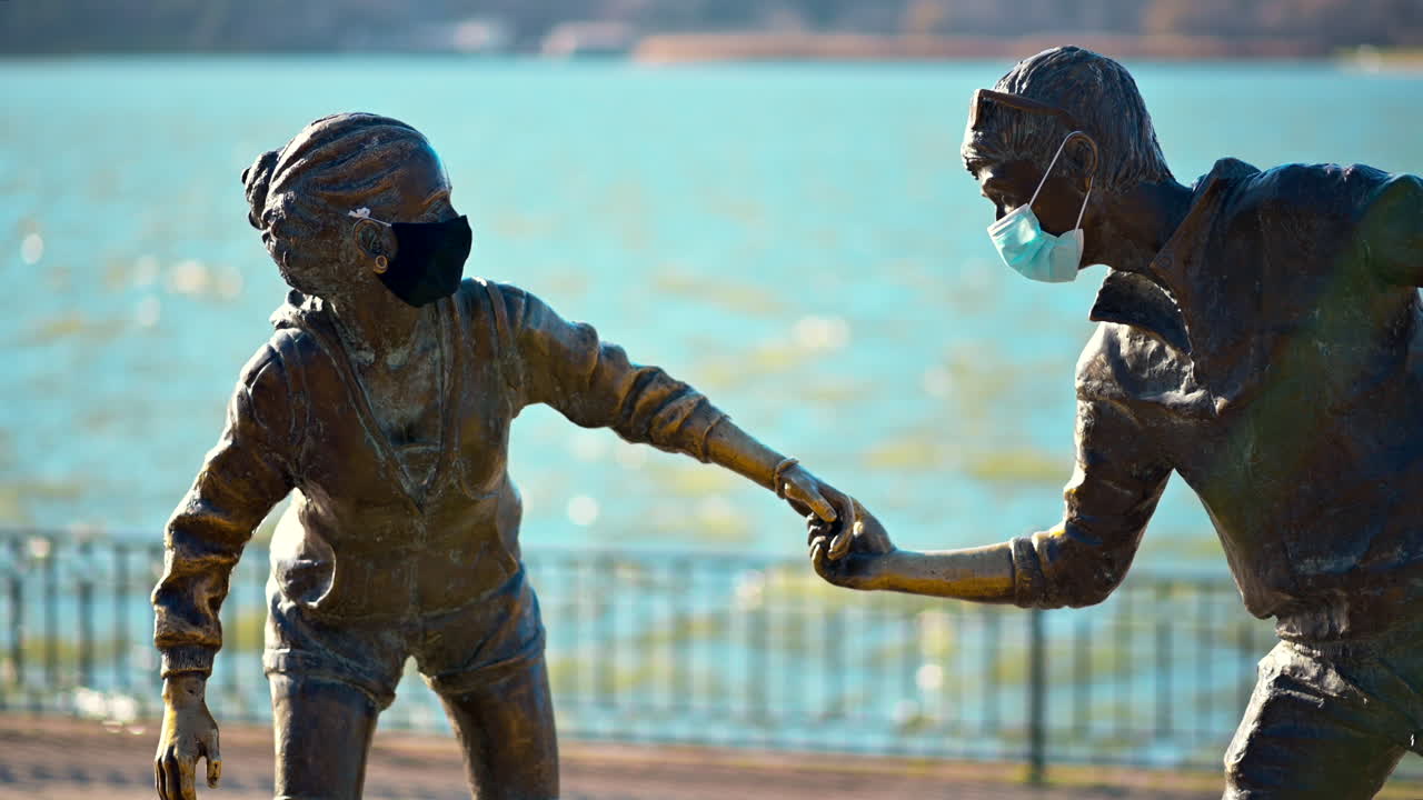 Two bronze sculptures of children hold hands and wear masks while standing by a lake. Sunlight glimmers on the water, creating a vibrant, lively atmosphere