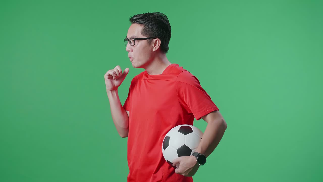 Vista laterale di un uomo asiatico eccitato con un pallone che fa il tifo per un tiro in porta mentre esulta per il calcio su sfondo green screen.