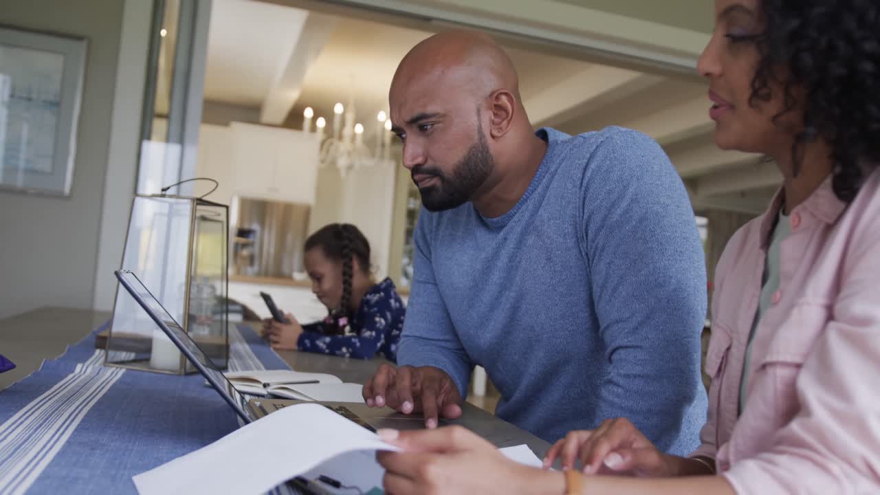 Happy biracial couple doing paperwork and using laptop at table, slow motion