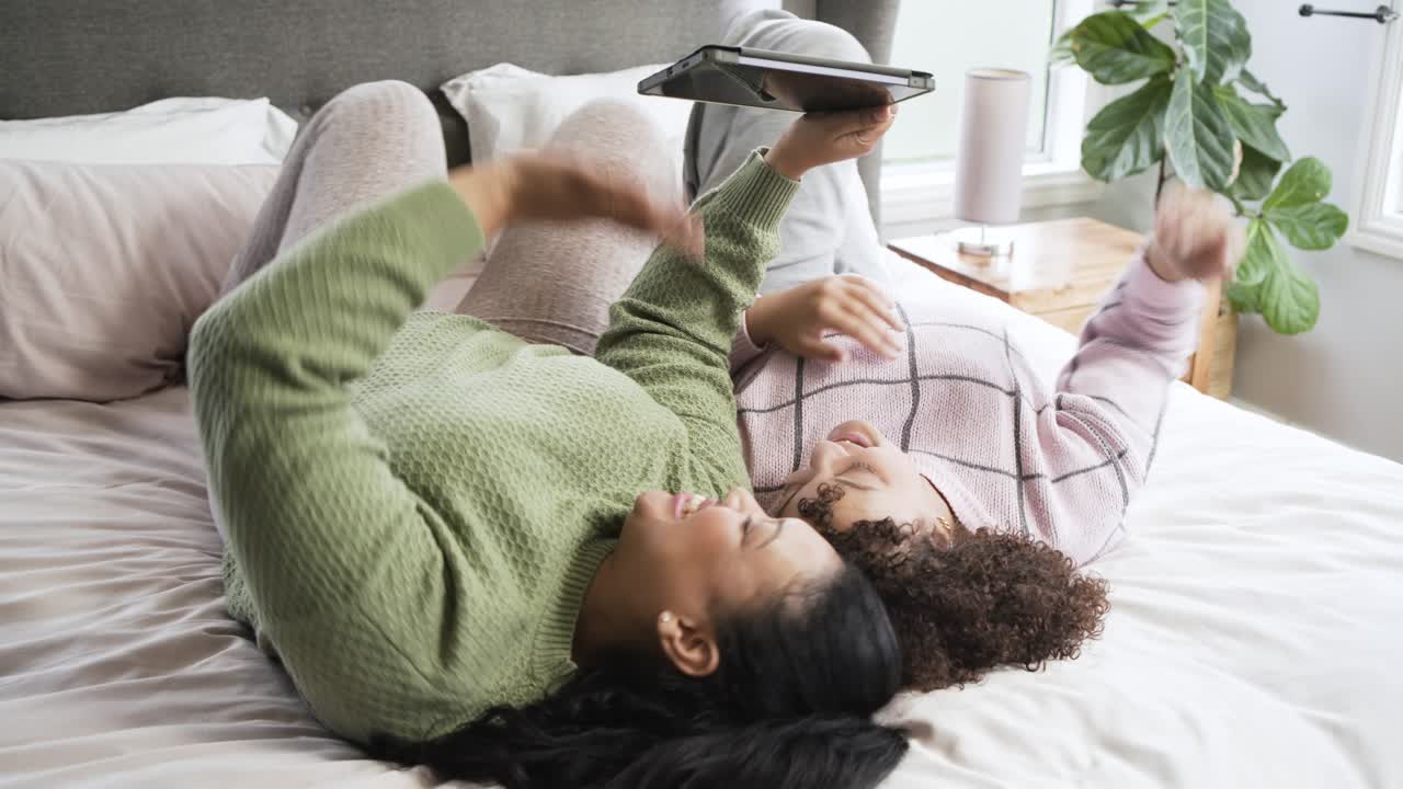 Madre e figlia sdraiate sul letto che cambiano l'angolazione del tablet per una vista migliore, allungandosi e scorrendo lo schermo.