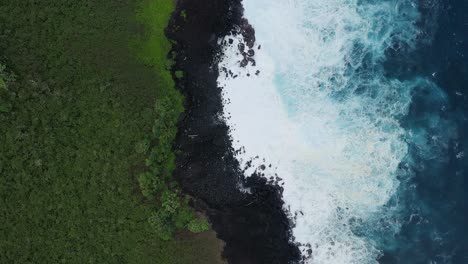 Aerial-view-of-waves-crashing-onto-a-rocky-Hawaiian-shoreline-with-lush-green-vegetation,-captured-in-dramatic-slow-motion