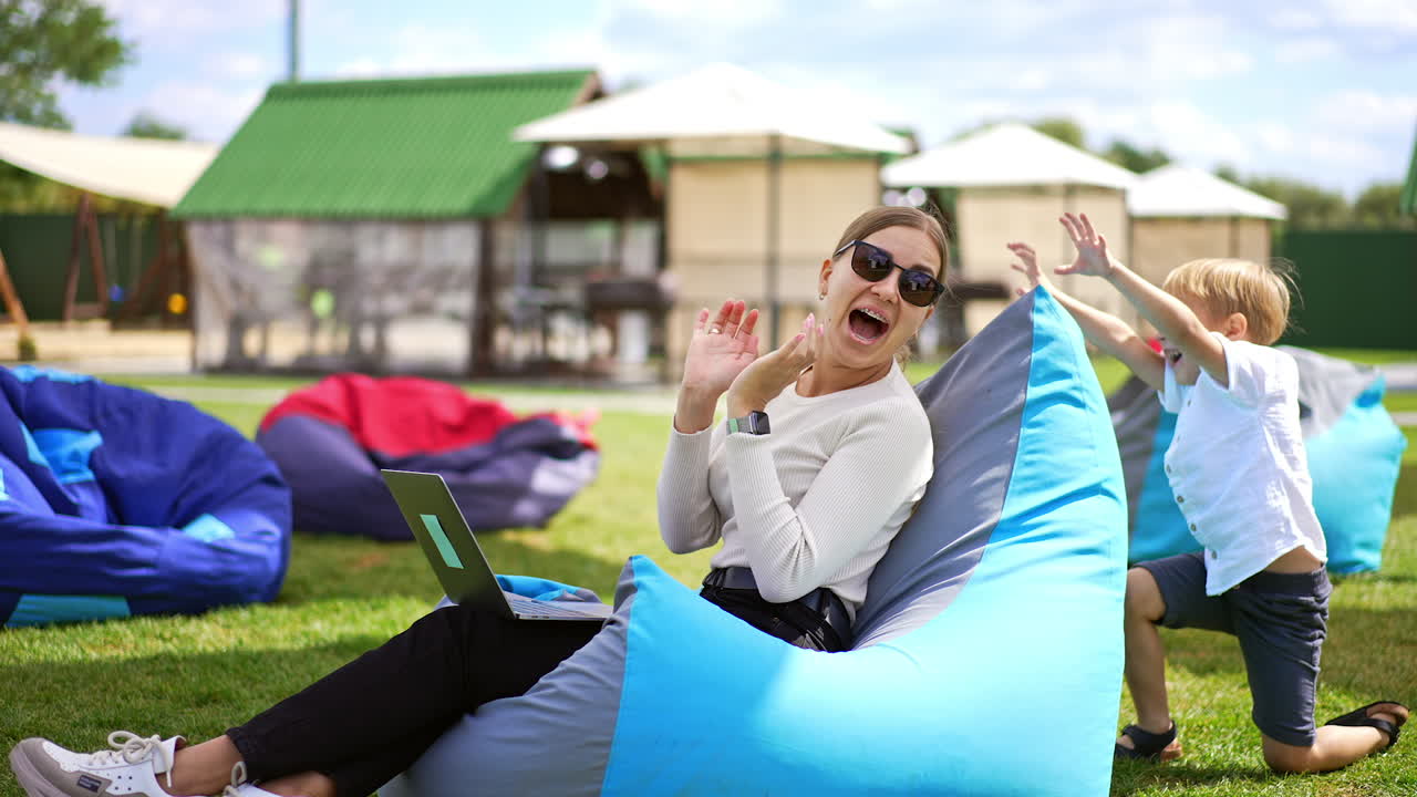 Long-haired woman working on laptop sitting in comfortable chair outdoors. Little boy comes up from behind trying to scare his mom. Playing with kid outside.