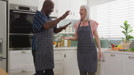feliz pareja diversa mayor usando delantales y bailando en la cocina