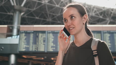 Parada-En-La-Pantalla-Con-Información-Sobre-La-Salida-Del-Avión-En-El-Aeropuerto,-Una-Joven-Con-Una-Mochila-Hablando-Por-Un-Teléfono-Móvil