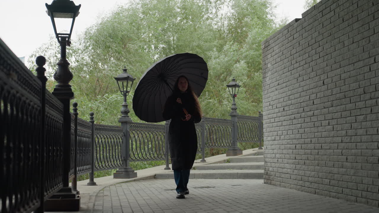 Donna che cammina con l'ombrello lungo una recinzione, lampioni vintage e sentiero acciottolato che creano un'atmosfera piovosa e malinconica, cappotto lungo e stivali, passeggiata solitaria di un pendolare, inquadratura cinematografica con verdi e grigi tenui
