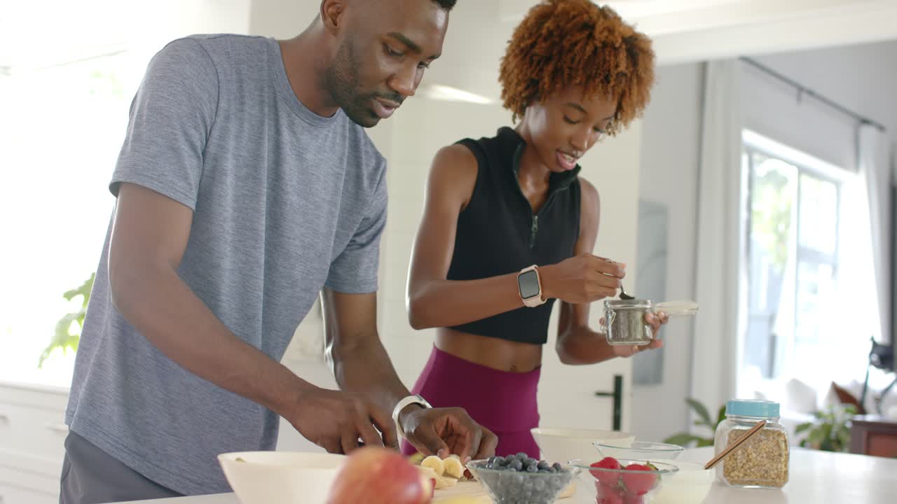 Afroamerikanisches Paar: Der Mann schneidet, an die Bar gelehnt, eine Banane, während die Frau aus einem Topf schöpft und Frühstück zubereitet.