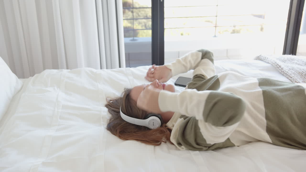 Relaxing on bed, young woman listening to music with headphones