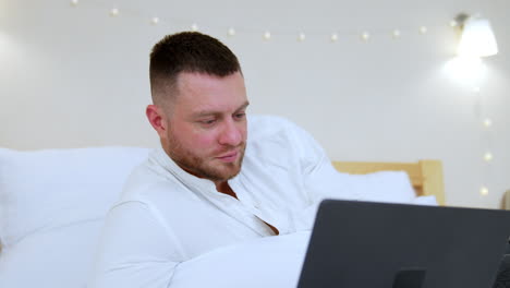 mid-aged caucasian man uses laptop lying on the bed. portrait of man working remote in comfortable atmosphere.
