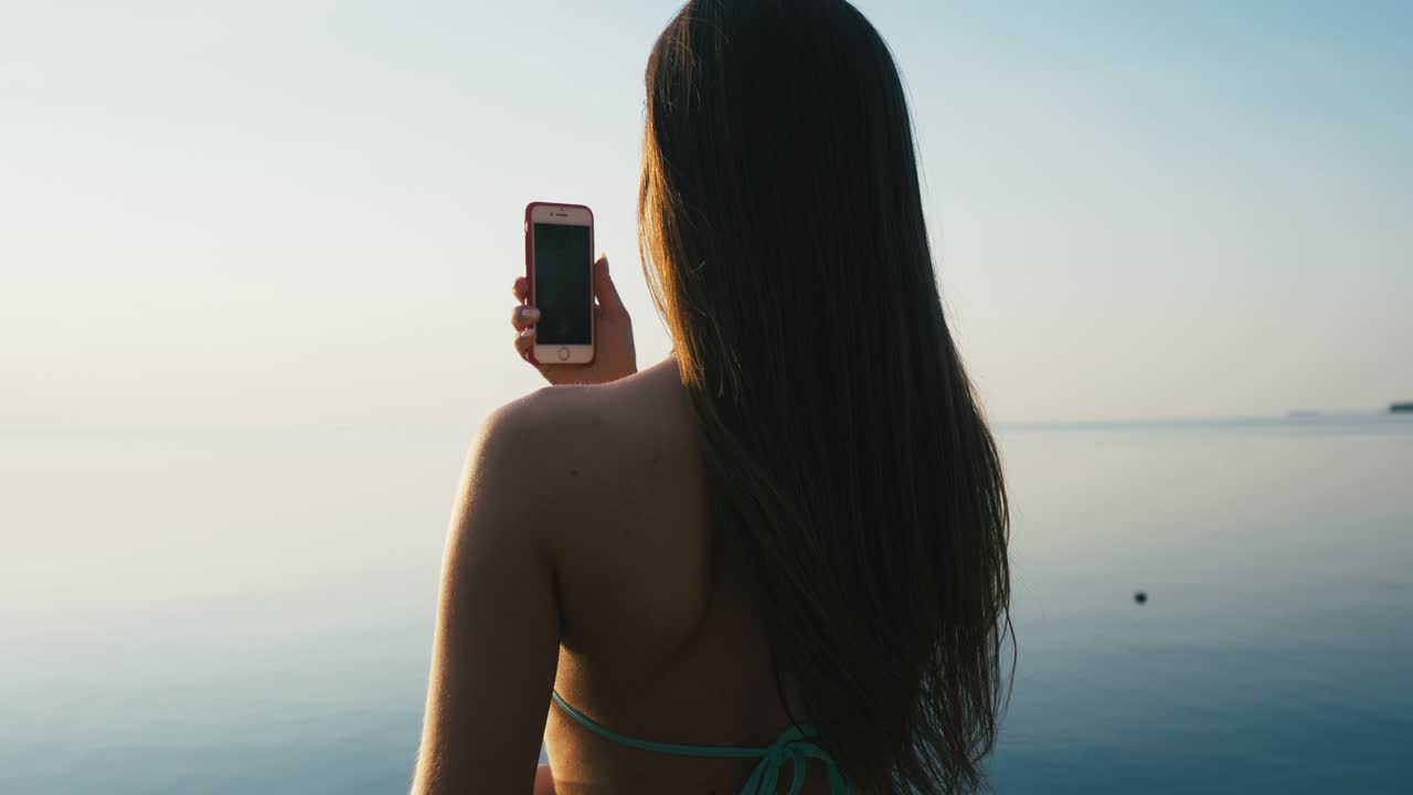 아름다운 젊은 여성이 휴가 중 해변 근처의 해가 지는 동안 스마트폰으로 사진을 찍고 있습니다.