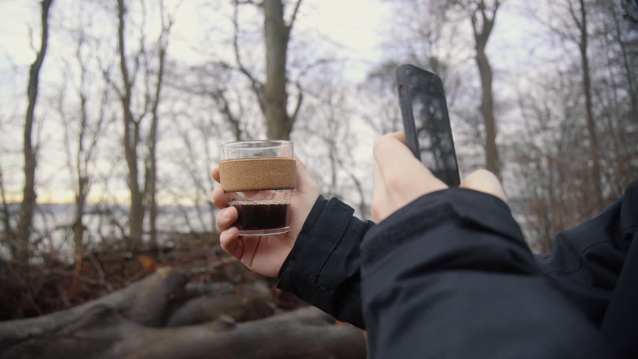 Young caucasian male taking a picture with his phone of hot coffee in a sustainable keep cup out of glass. Early in the morning outdoors forest at a lake