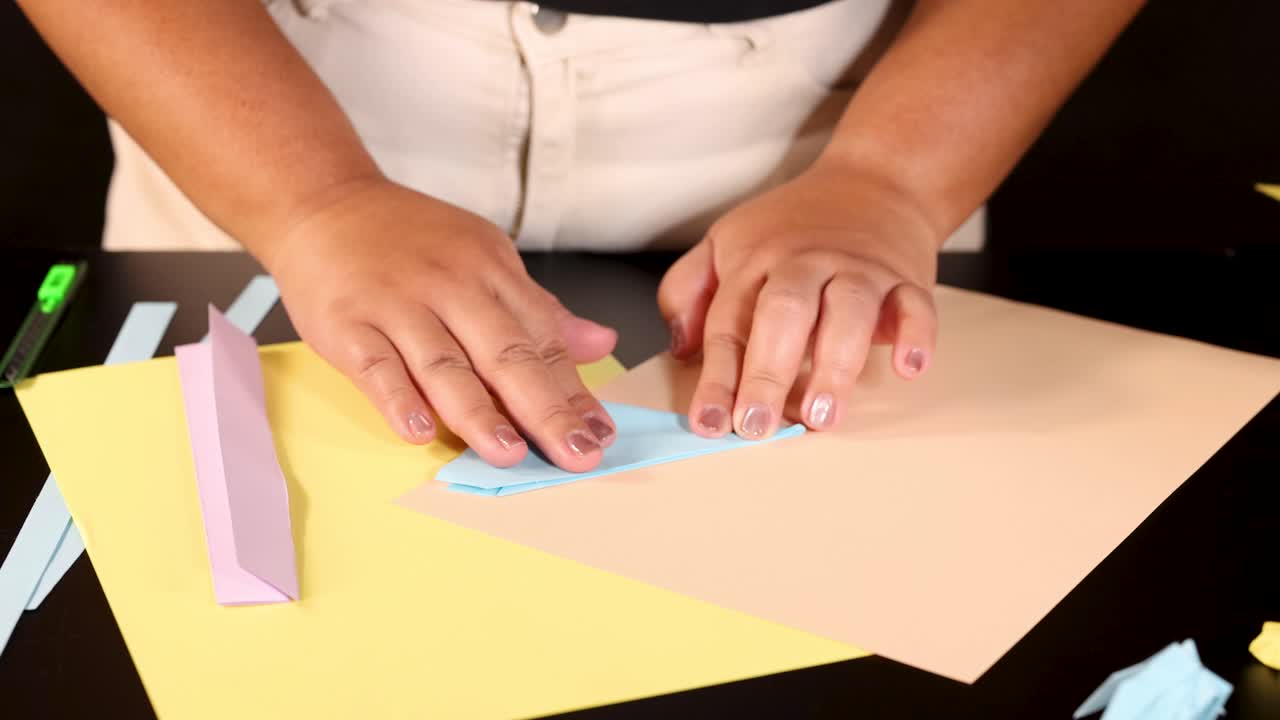 Person folds blue origami paper on desk under bright lighting, close-up, steady overhead camera
