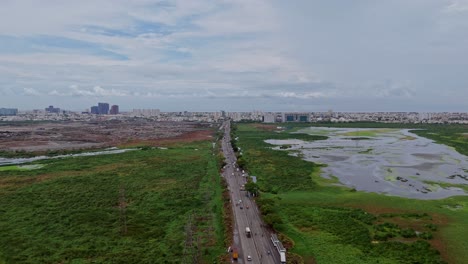 drone view of straight road cutting through green, marshy landsape with urban citysape at background.
