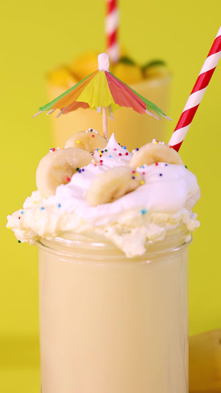Una mano decora un frullato di banana con un ombrellino da cocktail, panna montata e zuccherini su sfondo giallo