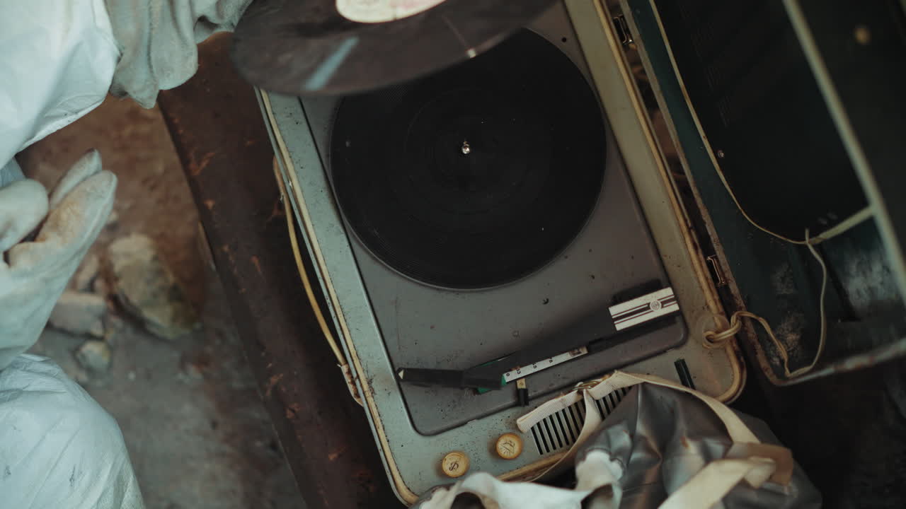 Aerial view of person wearing gloves carefully placing cartridge on worn vinyl record player with scratched surface, symbolizing nostalgia, music connection, and memory in difficult conditions