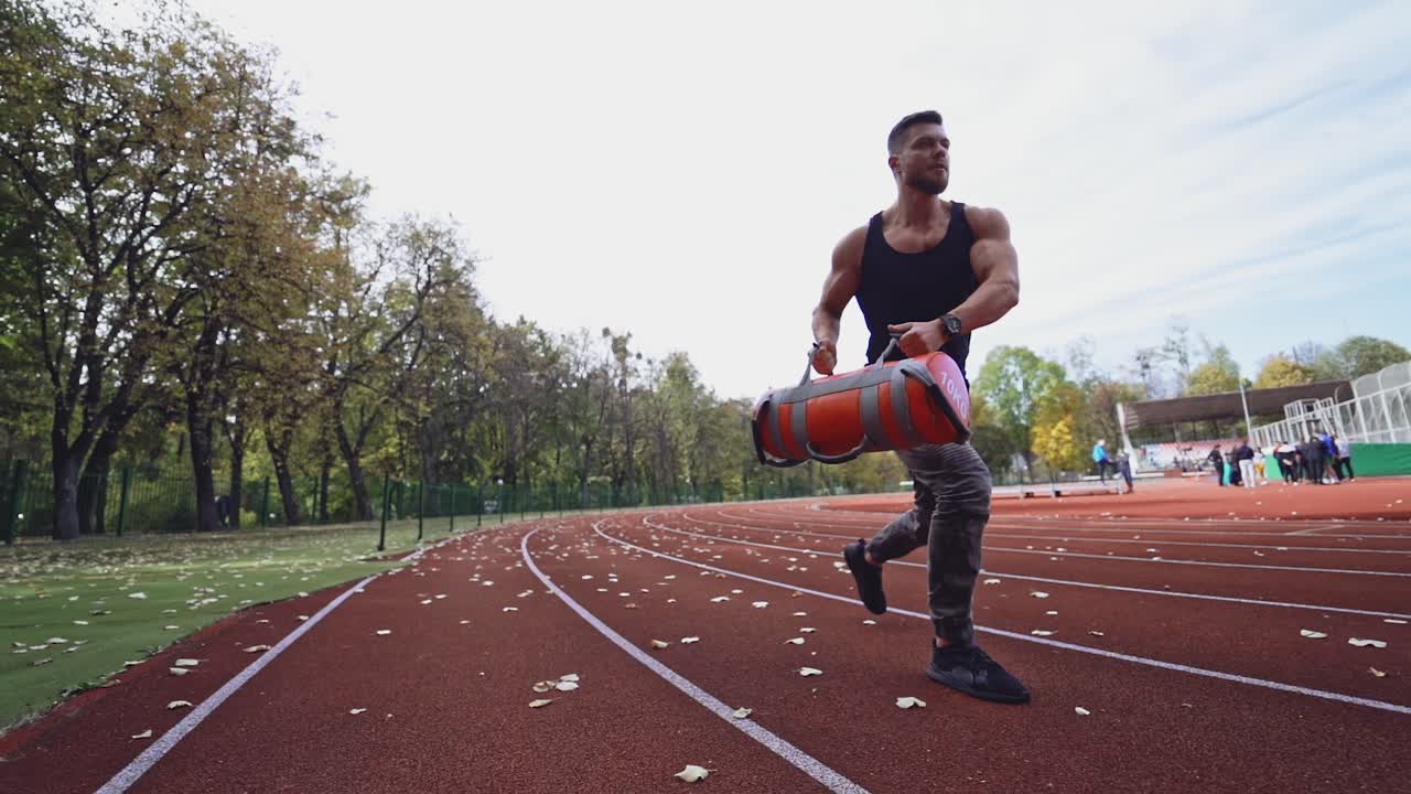 Strong athlete doing his workout on the stadium. Handsome muscular bodybuilder doing squats with weight outside. Slow motion.