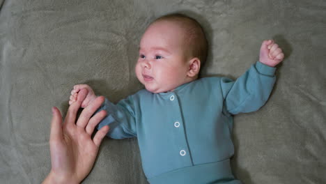 cute newborn baby in blue suit lies on bed holding parent's finger. beautiful little boy lies on his back on bed on grey background