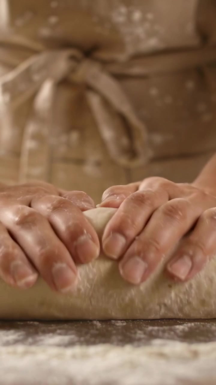 Vertical video: Shaping hands from dough ball on floury bench, apron tied, forming loaf to bake