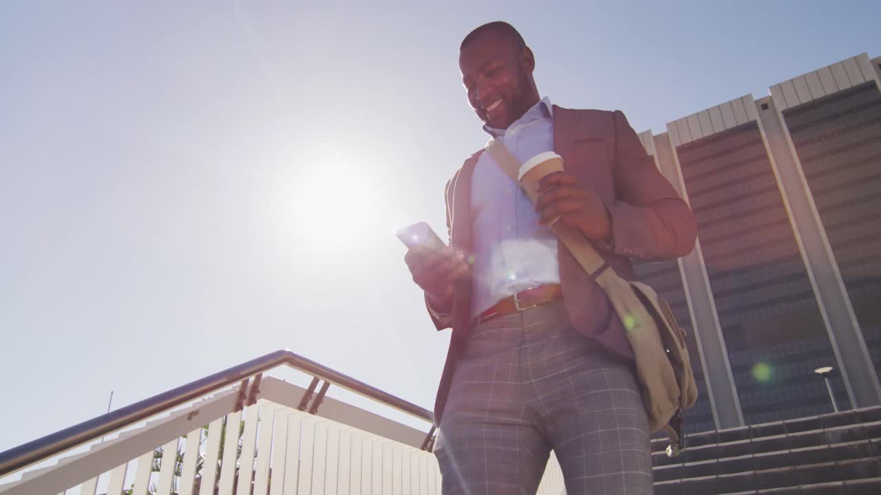 African american man in city walking downstairs, holding coffee, using smartphone