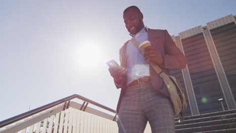 hombre afroamericano en la ciudad caminando por las escaleras, sosteniendo café, usando un teléfono inteligente