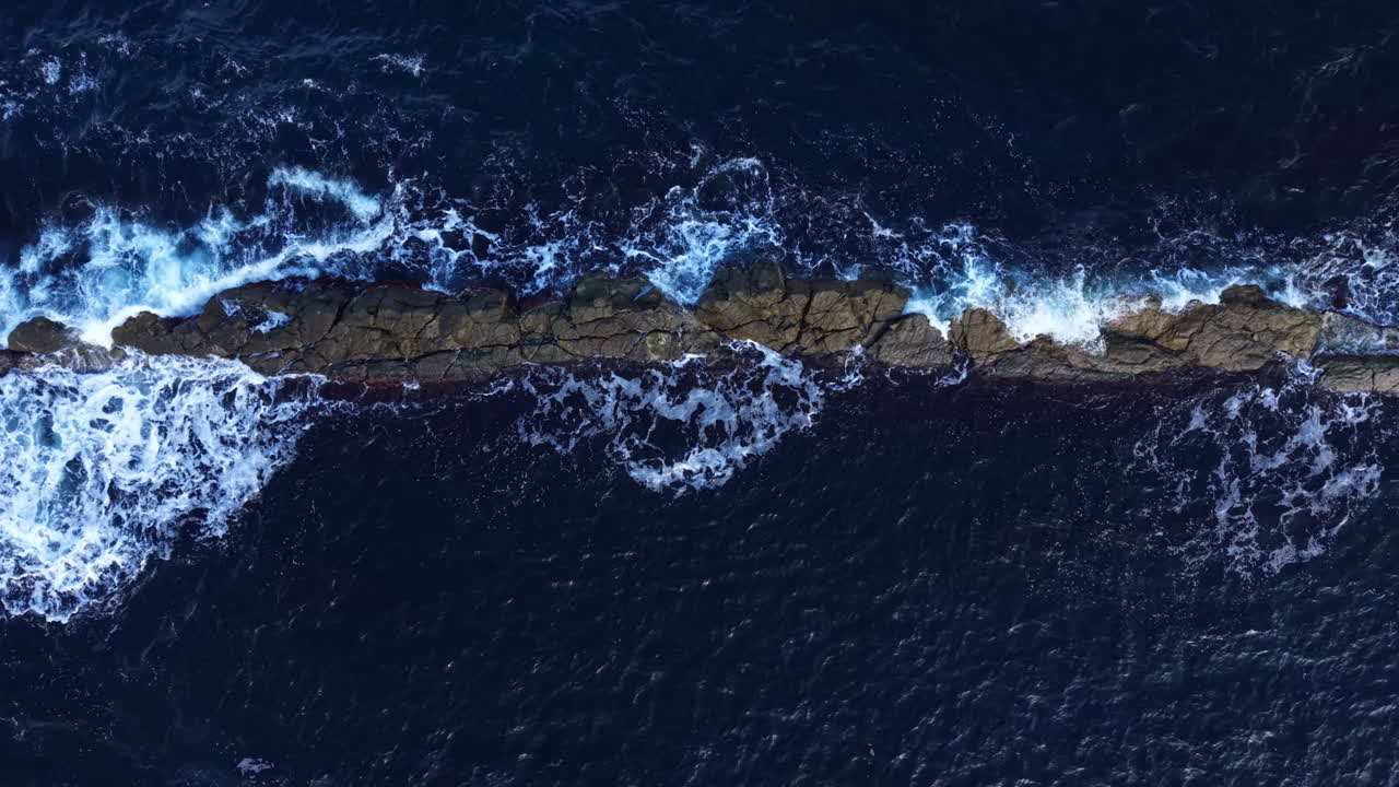 Waves crash over rocky outcrop under a bright sun amidst the endless blue ocean's expanse during a clear summer day