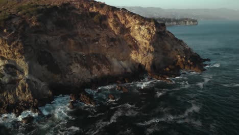 an aerial shot of the point dume cliffs in malibu in california as the waves crash against the rocks in the evening as the sunsets