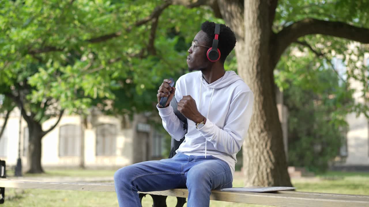 Portrait of joyful young man in headphones dancing sitting on bench at university yard outdoors. Happy carefree African American student enjoying hobby having fun in the morning. Education and joy.