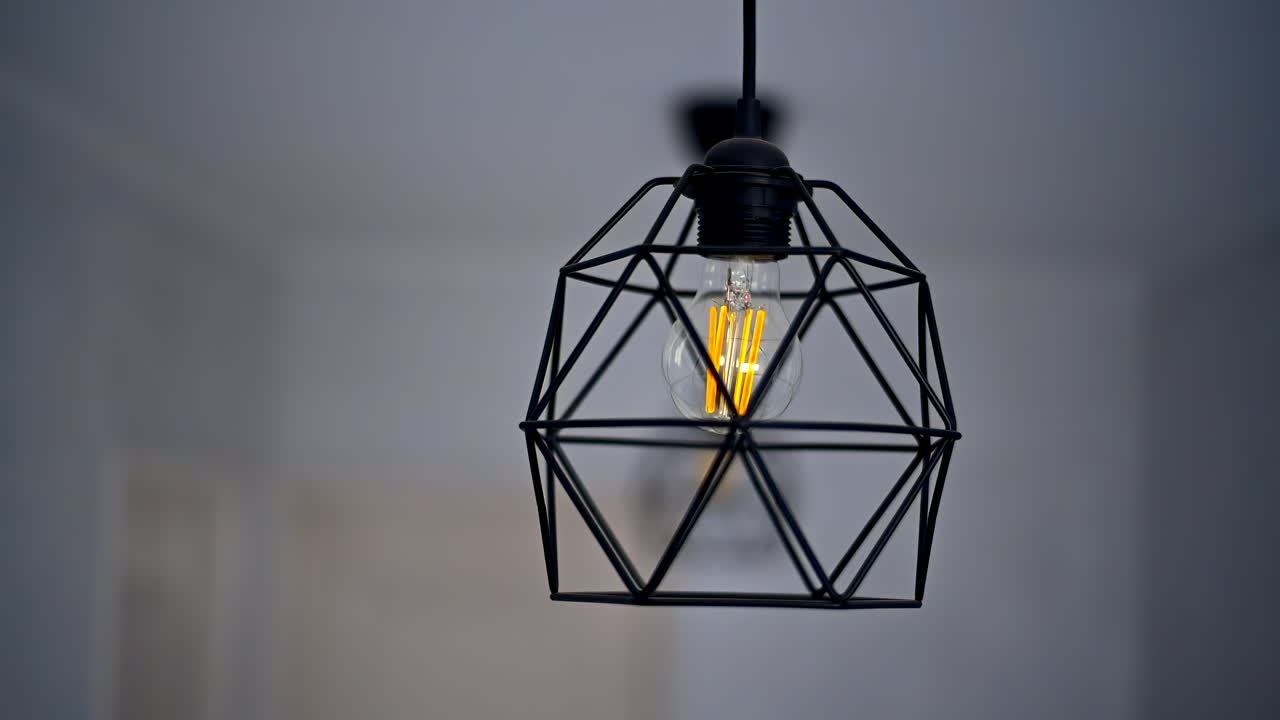 A stylish black geometric lamp hangs from the ceiling, casting a warm glow in a simple, uncluttered room with light walls and soft decor elements