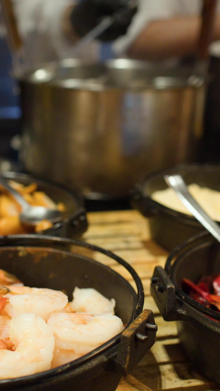 Una telecamera scorre su diverse pentole di ferro contenenti gamberetti, peperoncino e salse a un buffet.